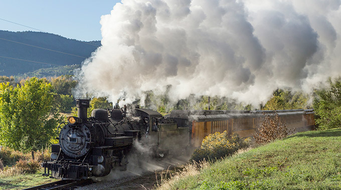 A steam engine train pulls passenger cars across a green landscape