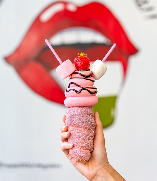 Someone holding up ice cream topped with marshmallows, a strawberry, and a chocolate drizzle, served in a dough cone